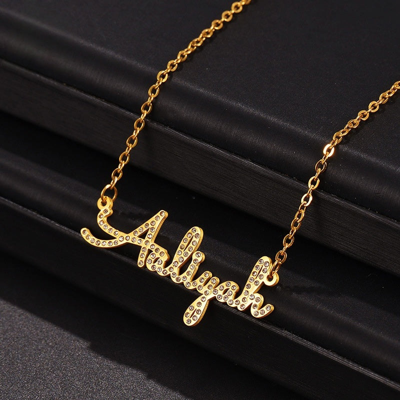 Gold stainless steel cursive name pendant fully paved with cubic zirconia, placed on black matte fabric for main product display